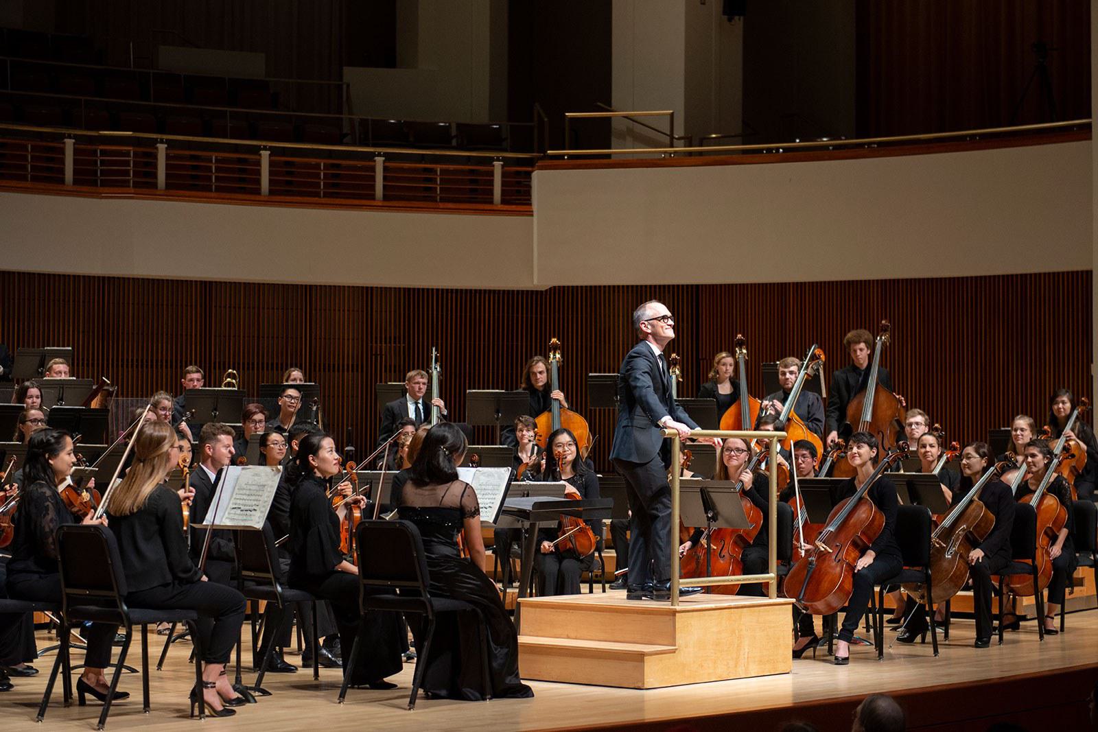 An orchestra and conductor perform on a concert hall stage.