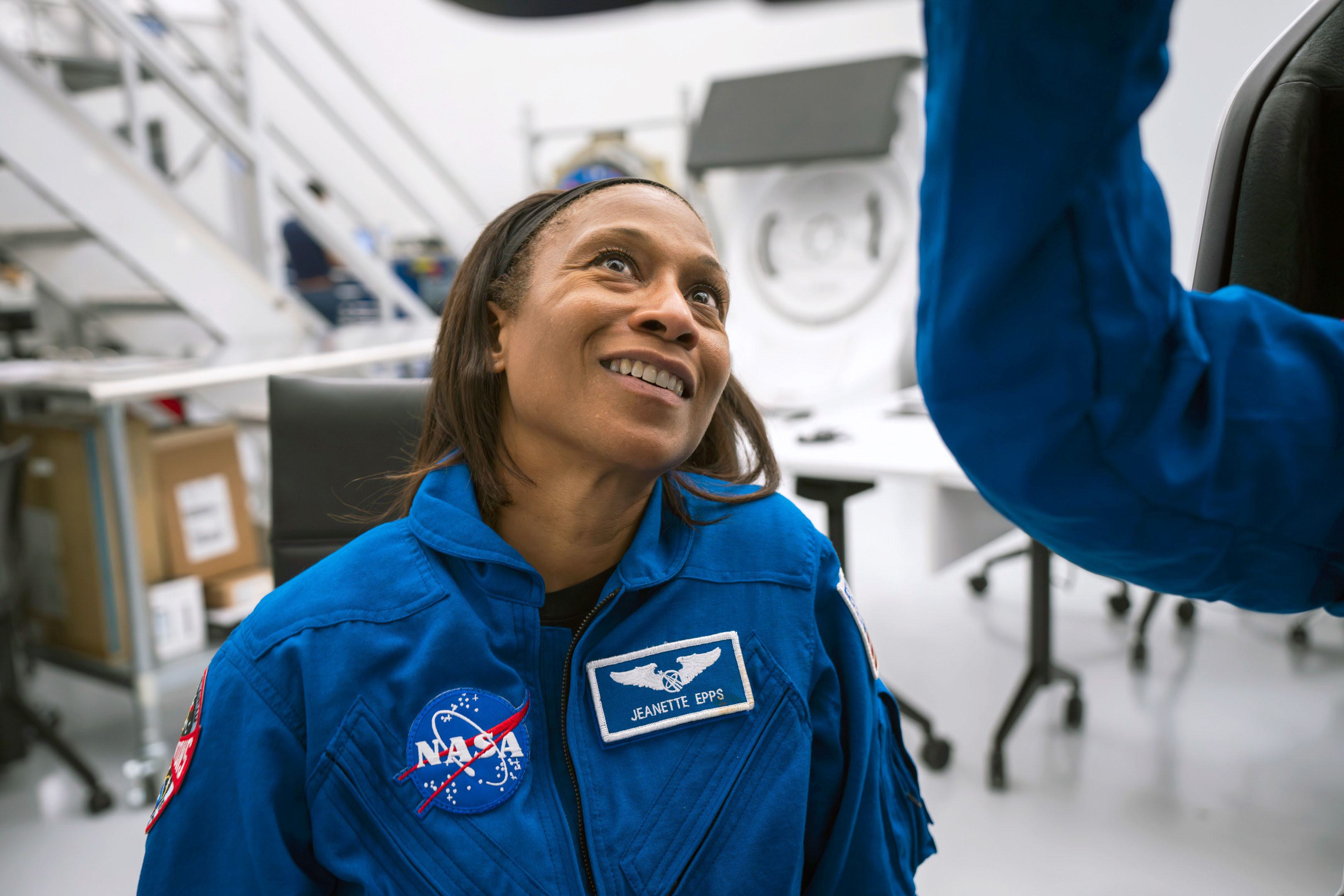 Jeanette Epps wearing a blue NASA jumpsuit with a stitched patch that reads "Jeanette Epps" on the left of her chest and the NASA logo on the right, smiling and looking up