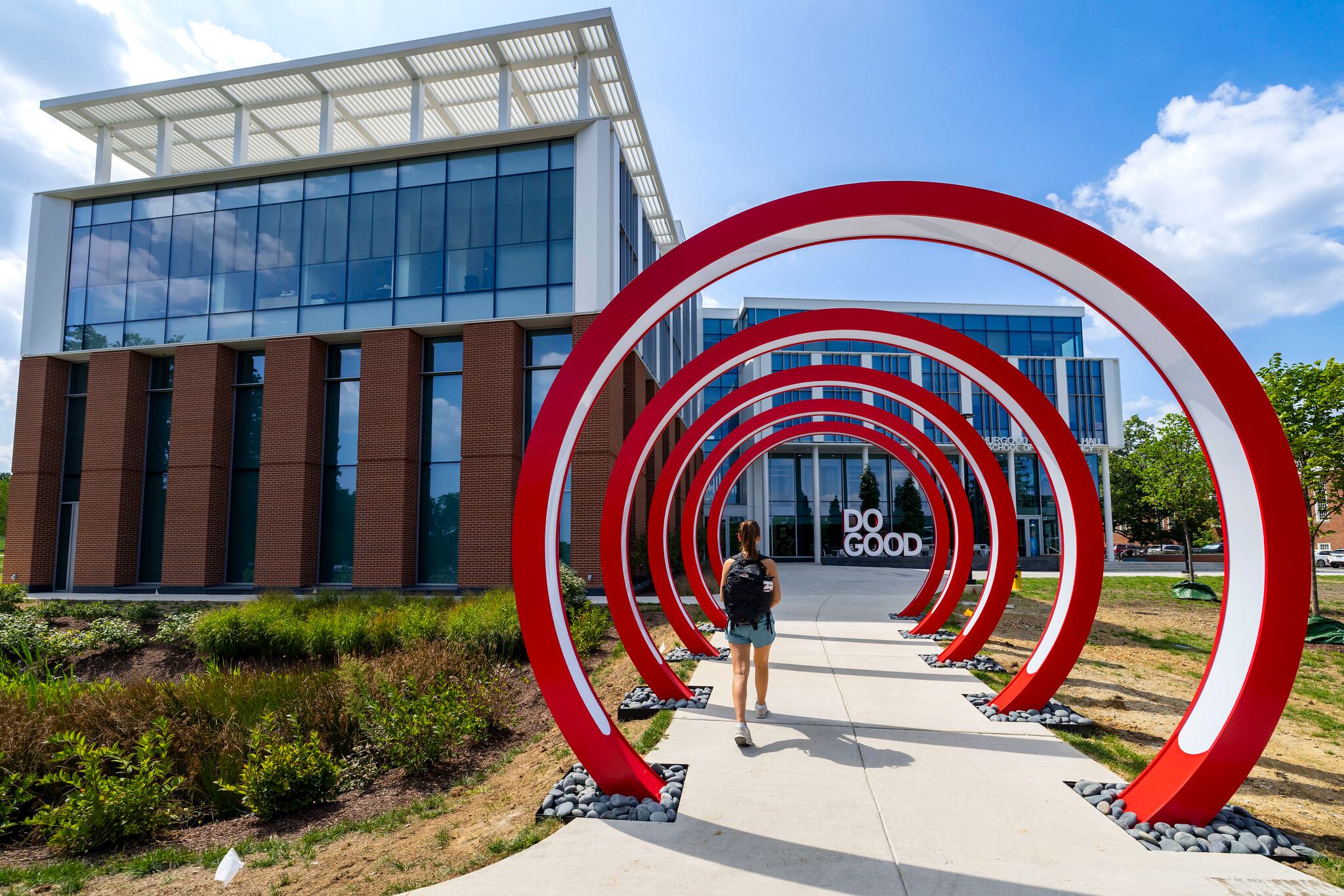 Female student walking through 6ft red rings to a glass building with a sculpture that says Do Good.