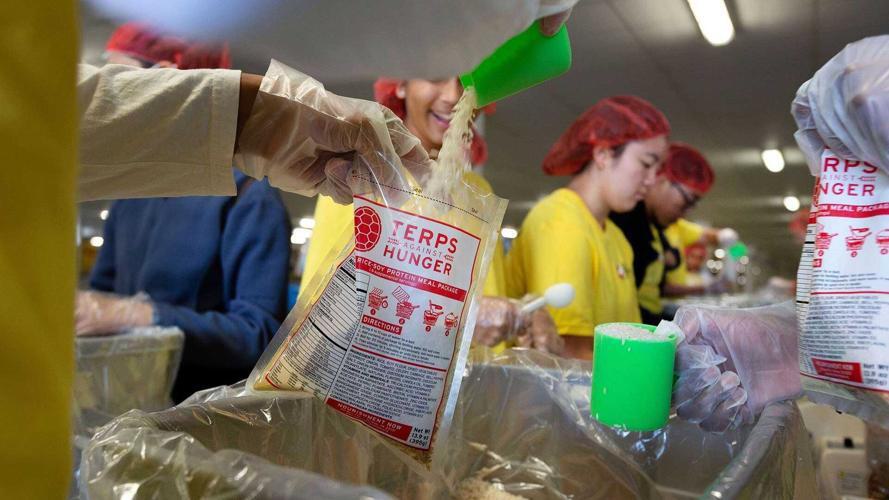 UMD Students are packing bags of food. In the foreground, a student is pouring beans into a TAH bag. In the background, students are wearing yellow shirts and hairnets.