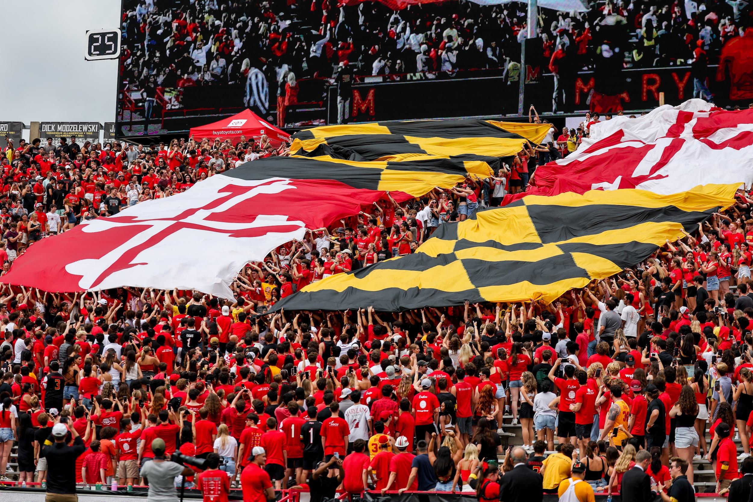 Image shows UMD fans dressed in red in stadium seating as the Maryland Flag unfurls above their heads