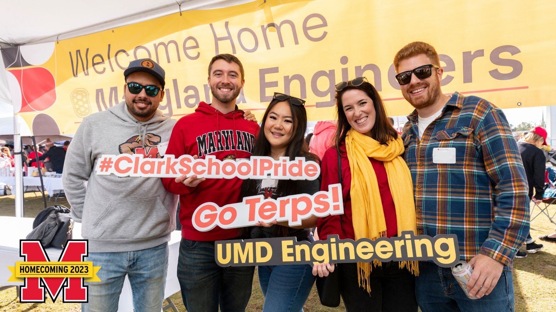 A group of young, smiling engineering alumni posing with photo props that read "#ClarkSchoolPride," "Go Terps," and "UMD Engineering" in bright UMD colors.