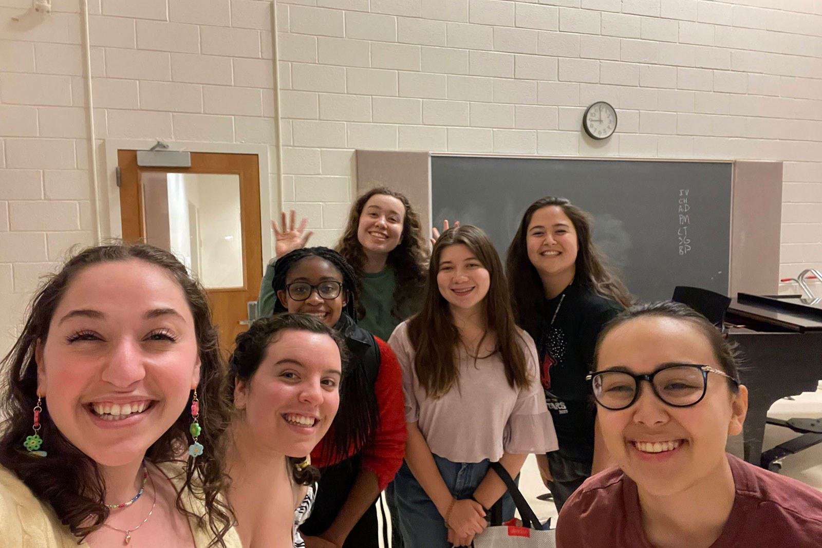 Seven women pose for a selfie together in a performing arts classroom.