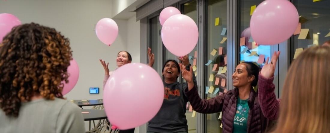Students keeping pink balloons afloat in a room lined with glass windows