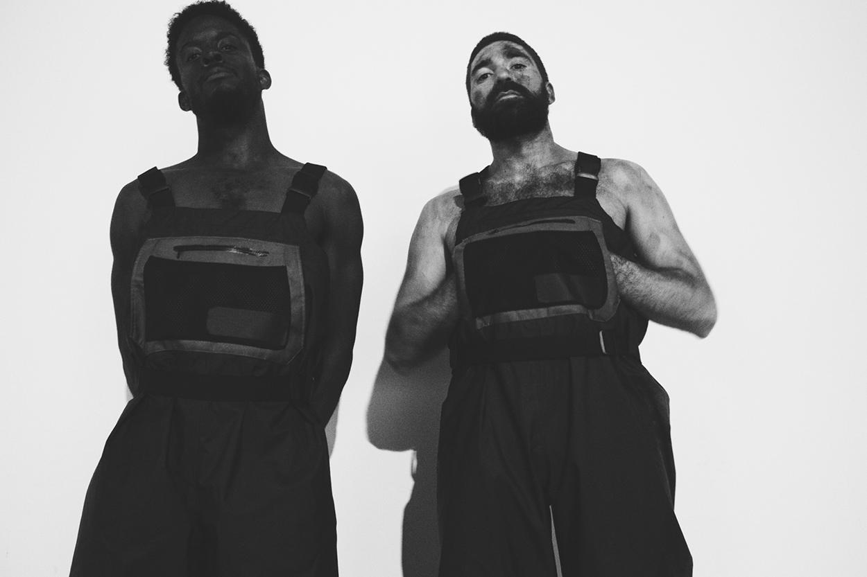 Two men pose in black jumpsuits.