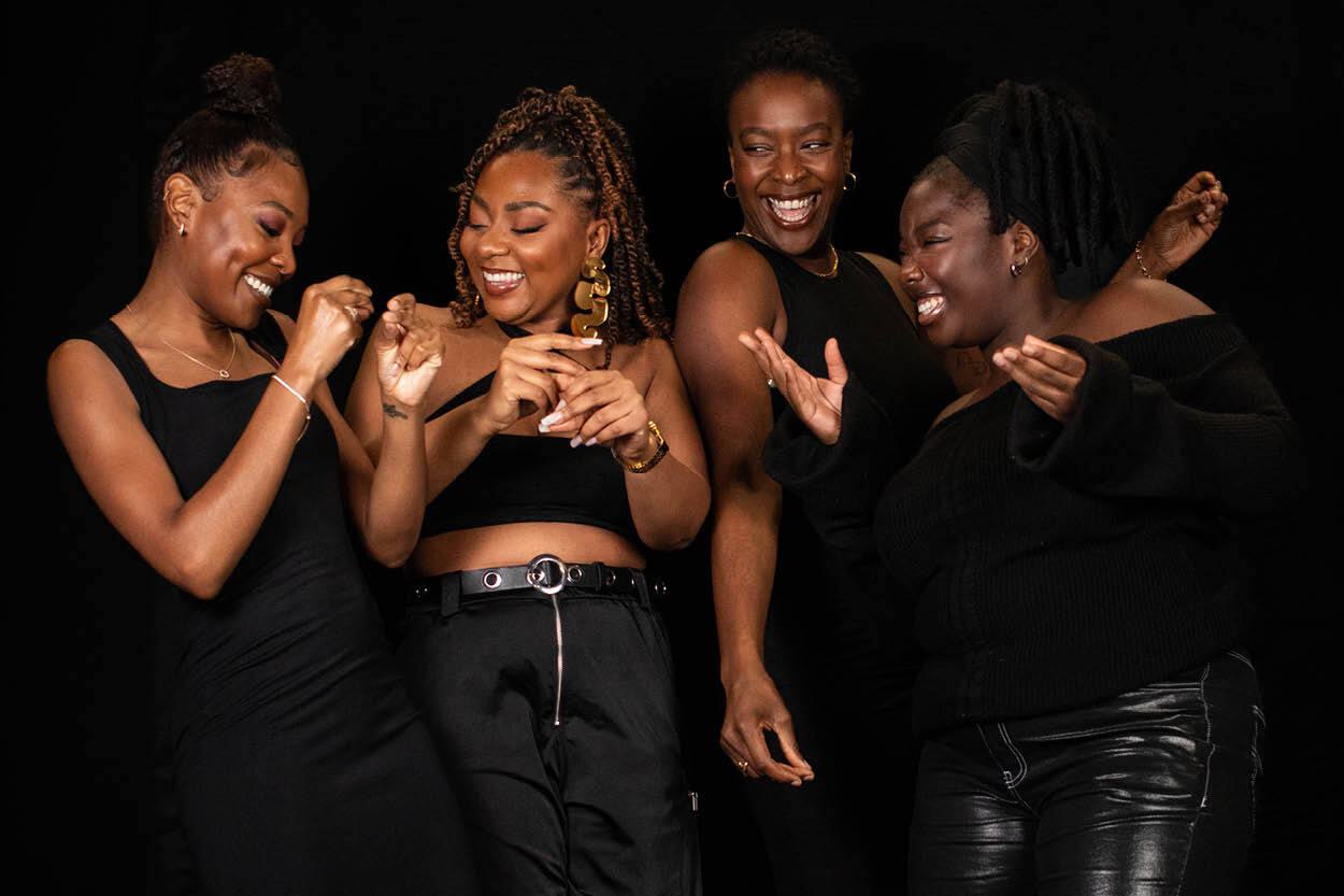 Four Black women laugh together against a black backdrop.