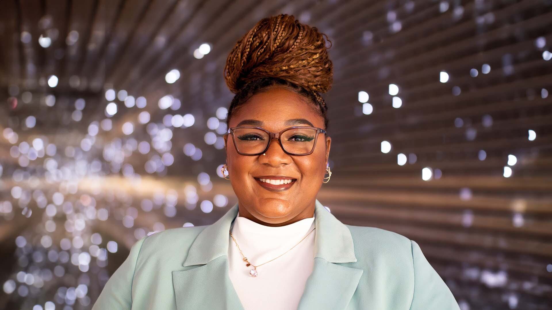 Tiara Fennell wearing a sea green blazer and white shirt. She is smiling and wearing brown glasses.