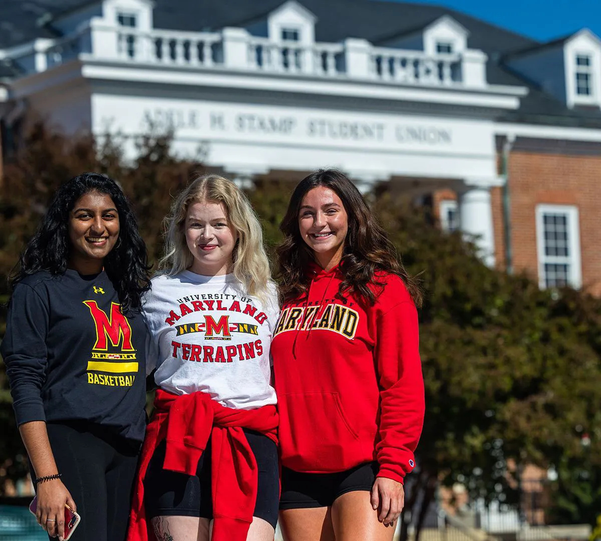 Three students pose in front of the Stamp Student Union in black, white and red Maryland gear.